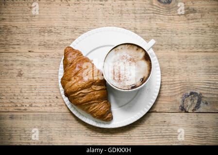 Tasse Latte Macchiato Kaffee mit Croissant auf weißen Teller auf alten rustikalen Holztisch angeordnet. Ansicht von oben Stockfoto