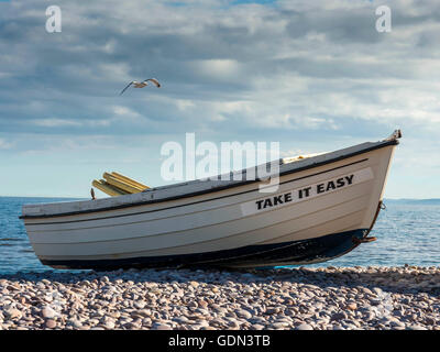 Take It Easy - Szene am Meer, mit einem blauen Ozean Hintergrund mit Boot in den mittleren Boden und einem Kiesel-Strand-Vordergrund. Stockfoto