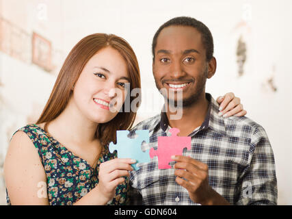 Interracial charmante Frau umarmen freundlich, große Puzzleteile hochhalten und glücklich Interaktion mit Spaß, verschwommen Studio-Hintergrund Stockfoto