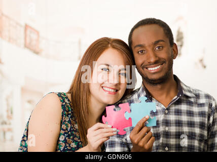 Interracial charmante Frau umarmen freundlich, große Puzzleteile hochhalten und glücklich Interaktion mit Spaß, verschwommen Studio-Hintergrund Stockfoto