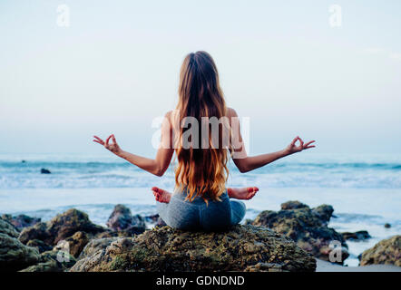 Rückansicht des junge Frau mit langen Haaren üben Lotus Yoga-Pose auf Felsen am Strand, Los Angeles, Kalifornien, USA Stockfoto