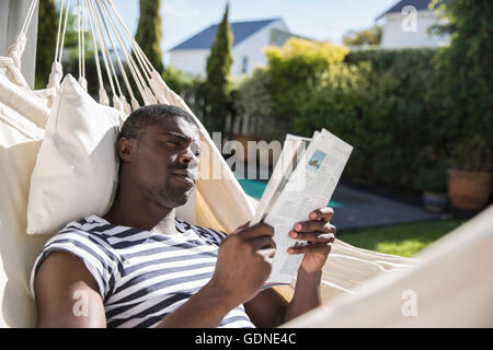 Junger Mann liegend auf Garten Hängematte Lesen einer Zeitschrift Stockfoto