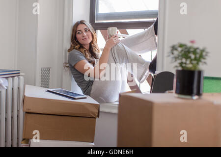 Junge Frau sitzt auf der Fensterbank der neue Heimat, Holding Heißgetränk, Karton Boxen im Zimmer Stockfoto