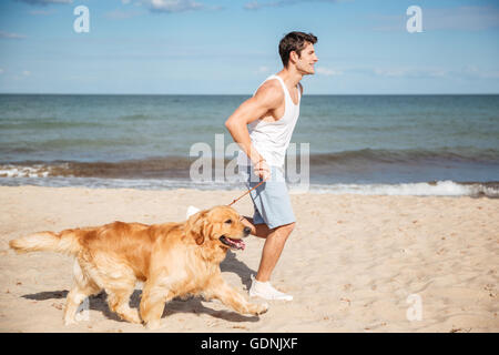 Hübscher junger Mann laufen und Spaß mit Hund am Strand Stockfoto