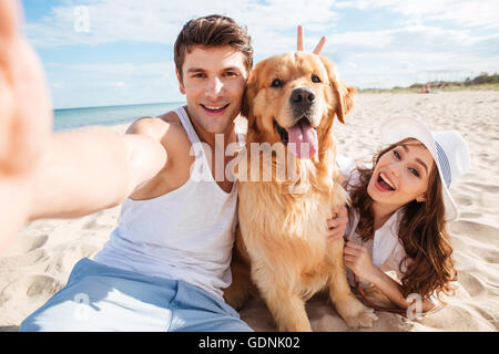 Junges glückliches Paar mit Hund unter einem Selfie am Strand Stockfoto