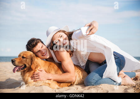 Lächelnd junges Paar mit Hund sprechen Selfie mit Smartphone am Strand Stockfoto