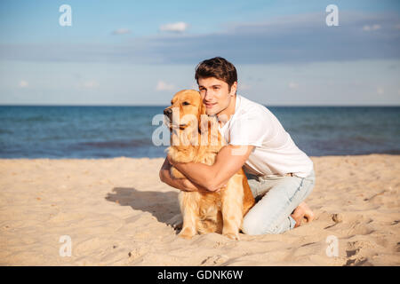 Hübscher junger Mann sitzen und umarmt seinen Hund am Strand Stockfoto