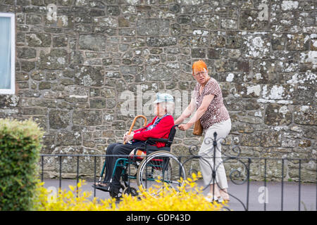 Ältere Frau im Rollstuhl geschoben von Frau mit orangen Haaren im Park spazieren, Shaftesbury, Dorset im Juli Stockfoto