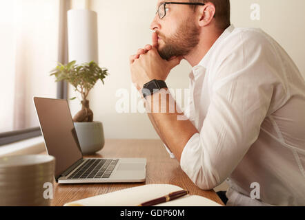 Seitenansicht der Geschäftsmann sitzt an seinem Schreibtisch mit einem Laptop und wegsehen. Mann an seinem Workdesk denken von Business-Lösungen. Stockfoto