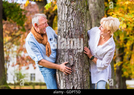 Paar, senior Mann und Frau, flirten mit einander spielen verstecken und suchen, um einen Baum im Herbst Baum Stockfoto
