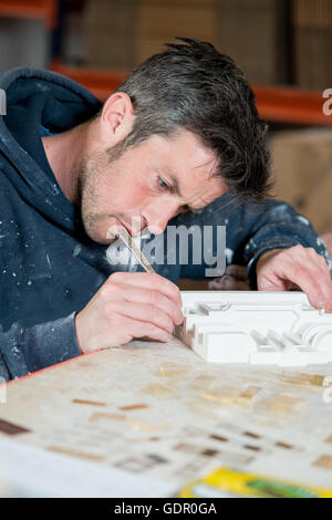 Ein Kaukasischen Männchen in Farbe bespritzt-Hoodie mit einer Präzision Messer und schneiden einen Abschnitt eines weißen Putz-Modells auf einem Schreibtisch. Stockfoto