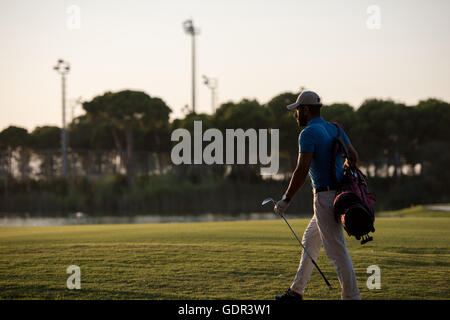schöne orientalische Golfer Tragetasche und zu Fuß zum nächsten Loch am Golfplatz auf wunderschönen Sonnenuntergang im Hintergrund Stockfoto