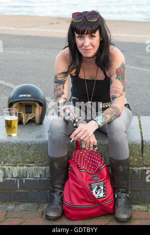 Junge Frau mit Tattoos sitzen auf Wand mit Glas trinken und Helm auf Traum Maschinen Fahrrad Nacht am Poole Quay, Dorset Stockfoto