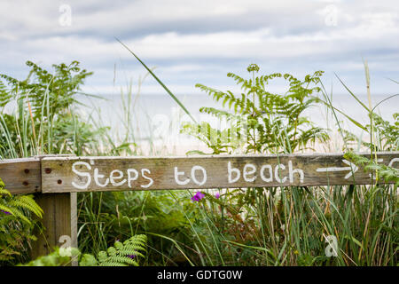 Weiß lackierter Schild mit der Aufschrift, dass "Schritte zum Strand" Pfeil auf Sonne gebleicht Holzzaun mit Blick auf den Strand und das Meer hinaus. Stockfoto