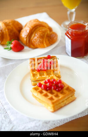 Waffeln mit Johannisbeer-Marmelade und Beeren auf einem weißen Teller, Croissants, Orangensaft auf dem hölzernen Hintergrund Stockfoto