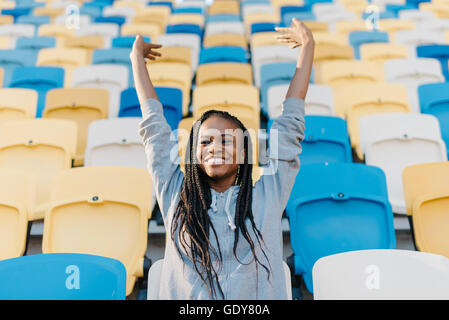 Glücklich afroamerikanische Frau stehend in unter den leeren Sitzreihen im Stadion anfeuern mit ihren Armen hob Stanz Luft Stockfoto