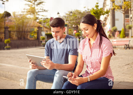 Süße junge Paar sitzen in einem Stadtplatz ruht beim Betrachten ihrer Tablet und Handy während lässig in Jeans gekleidet Stockfoto
