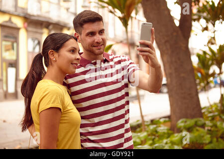 Junges Paar Blick auf das Handy glücklich lächelnd, während der Einnahme einer Selfies bekleidet lässig T-shirts in einer von Bäumen gesäumten Straße Stockfoto