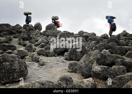 Drei Träger tragen große Taschen auf dem Kopf und großen vulkanischen Felsen, Mount Kilimanjaro National Park, Tansania Stockfoto