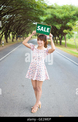 Nette Frau Hand mit grüner Grundplatte Schild mit Text "Verkauf" auf Straße und Baum, Smiling weibliches Modell. Stockfoto