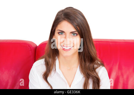 Porträt der jungen Frau lächelnd. Hübsche Frau mit langen braunen Haaren sitzt auf roten Sofa. Sie ist zuversichtlich Blick auf er Stockfoto