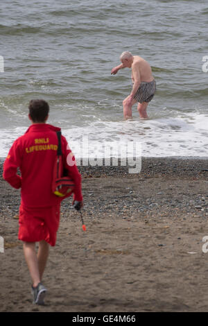 Ein RNLI Rettungsschwimmer stehen auf Aberystwyth Strand beobachten im Auge zu behalten, auf einen leitenden Erwachsenen männlichen Mann Schwimmen im Meer, UK Stockfoto