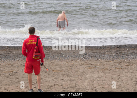 Ein RNLI Rettungsschwimmer stehen auf Aberystwyth Strand beobachten im Auge zu behalten, auf einen leitenden Erwachsenen männlichen Mann Schwimmen im Meer, UK Stockfoto