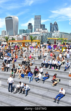 Büromitarbeiter machen Mittagspause bei heißem Sonnenschein im Amphitheater More London Scoop am Flussufer mit der Skyline der City of London jenseits von Southwark England UK Stockfoto