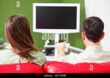 Heckansicht des jungen Paares vor dem Fernseher. Männliche und weibliche Partner sitzen auf roten Sofa. Sie sind zu Hause. Stockfoto