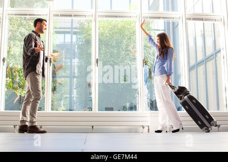 Voller Länge Frau mit Gepäck zu ihrem Freund winken. Junge männliche und weibliche Partner sind am Flughafen. Sie sind in Casuals. Stockfoto