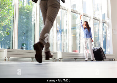 Glückliche junge Frau mit Gepäck zu ihrem Freund winken. Mann läuft in Richtung Partnerin am Flughafen. Sie ist mit Gepäck an de Stockfoto