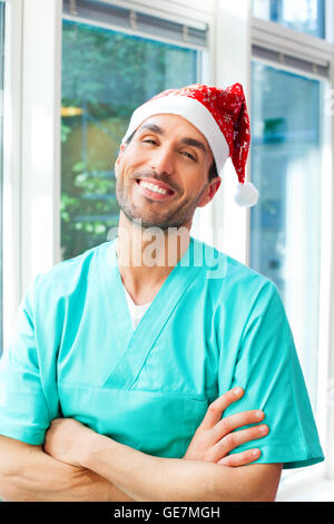 Porträt von glücklichen männlichen Chirurg Santa Hut trägt. Junger Arzt steht mit verschränkten Armen. Er trägt Peelings Stockfoto