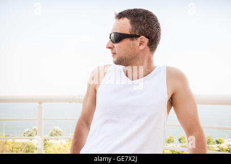 Junge sportliche kaukasischen Mann im weißen Hemd und Sonnenbrille. Outdoor-Sommer-Porträt mit Meerblick auf einem Hintergrund Stockfoto