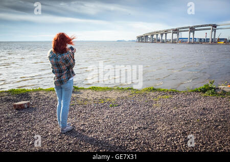 Teenager-Mädchen mit roten Haaren unter Telefon Landschaftsfoto mit modernen Brücke im Bau auf einer Küste Stockfoto