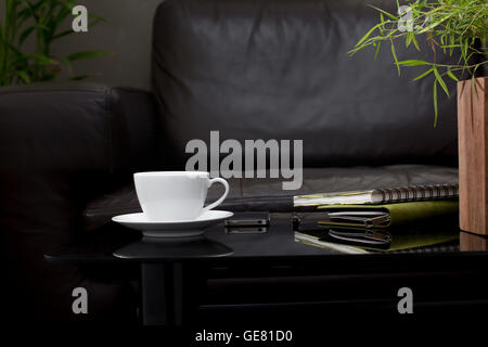 weiße Kaffeetasse auf Glastisch mit dunklen braunen Sofa im Wohnzimmer Stockfoto