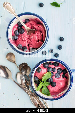 Hausgemachte Heidelbeer-Eis schöpft mit frischen Beeren und Minze in Emaille Becher über hellblauen Hintergrund Stockfoto