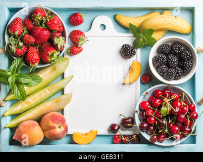 Gesunden Sommer Obstsorte. Süße Kirschen, Erdbeeren, Brombeeren, Pfirsiche, Bananen, Melonenscheiben und Minze Blätter auf blauen Hintergrund mit weißer Keramik Board in Mitte Stockfoto