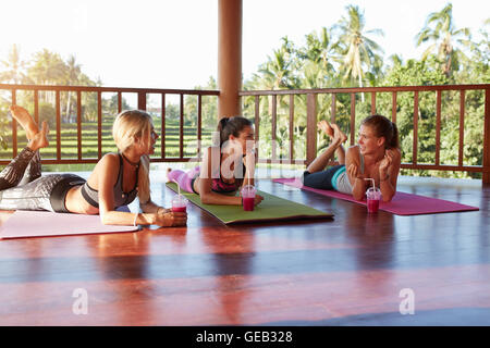 Drei schöne Mädchen mit Saft aus frischen Früchten auf dem Boden liegend und im Chat nach Yoga zu praktizieren. Gruppe von Frauen rela Stockfoto