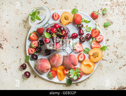 Gesunden Sommer Obstsorte. Süße Kirschen, Erdbeeren, Pfirsiche, Aprikosen und Minze Blätter auf weißer Keramik Teller über leichte konkrete Hintergrund Stockfoto