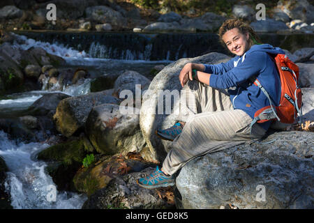 Nette junge Frau sitzen auf Stein in der Nähe von Mountain Creek lächelnd und genießen Sie warme Sonnenlicht weibliche Wanderer mit Rucksack und sportlichen C Stockfoto