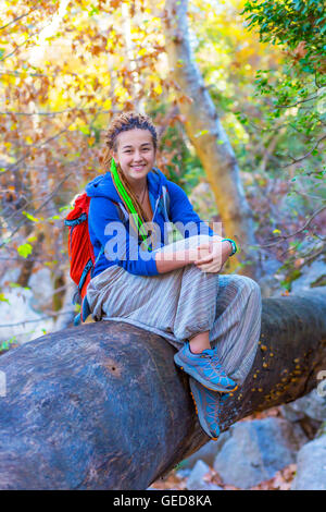 Fröhliche Mädchen mit Rucksack und Hippie-Stil Kleidung sitzen auf großen Stamm von einem umgestürzten Baum im Wald mit Sonnenstrahlen kommenden thro Stockfoto