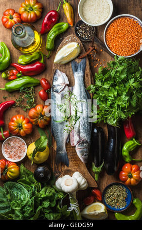 Rohe ungekochte Wolfsbarsch Fisch, Gemüse, Getreide, Kräuter, Reis Gewürzen und Olivenöl auf rustikale Schneidbrett aus Holz, Ansicht von oben Stockfoto