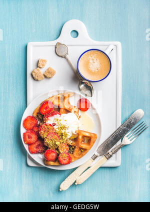 Frühstücks-Set. Warme hausgemachte belgischen Waffeln mit Sahne, Erdbeeren, Ahornsirup, zerkleinerten Pistazien, Espresso und braunem Zucker auf weißer Keramik Board über blau lackierten hölzernen Hintergrund Stockfoto