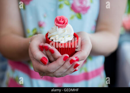 Schuss ein Womans Hände halten einen hübschen Cupcake hautnah. Sie trägt ein helles Blumenkleid Deisgn. Der Cupcake ist in einem roten p Stockfoto