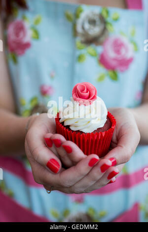 Schuss ein Womans Hände halten einen hübschen Cupcake hautnah. Sie trägt ein helles Blumenkleid Deisgn. Der Cupcake ist in einem roten p Stockfoto