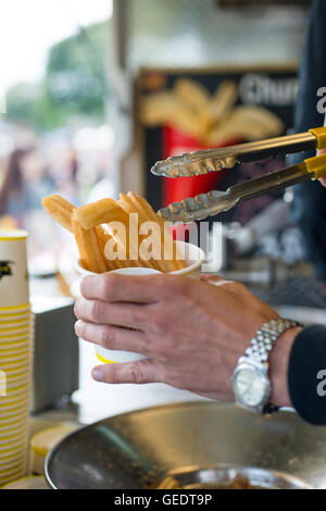 Nahaufnahme eines Mannes halten Metall Zange dienen tief gebratene süße Churros in einen Pappbecher in eine Outdoor-stall Stockfoto