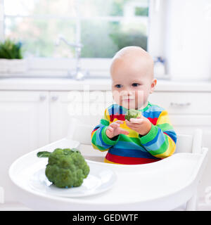 Glückliches Baby sitzen im Hochstuhl Essen Brokkoli in eine weiße Küche. Gesunde Ernährung für Kinder. Bio-Gemüse als Beikost Stockfoto
