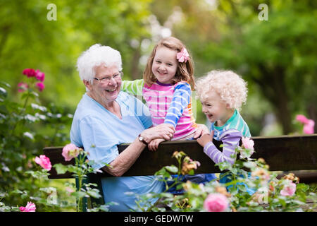 Glücklich senior Lady spielt mit kleinen Jungen und Mädchen in blühender Rosengarten. Großmutter mit Enkel sitzen auf einer Bank Stockfoto