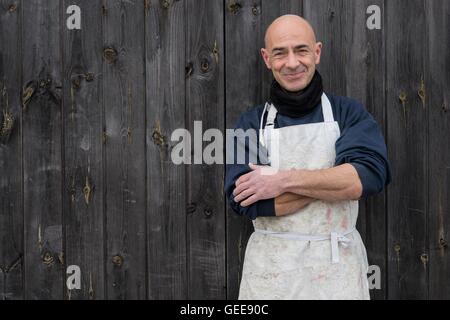 Kahlköpfiger Mann in dicke Kleidung und weiße Schürze lächelnd und mit verschränkten in eine Holzplatte Hintergrund stehend Stockfoto
