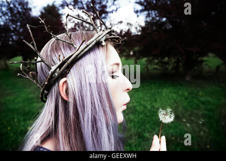 Junge Frau mit lila Haaren Zweig Krone trägt und bläst auf einem Löwenzahn Stockfoto